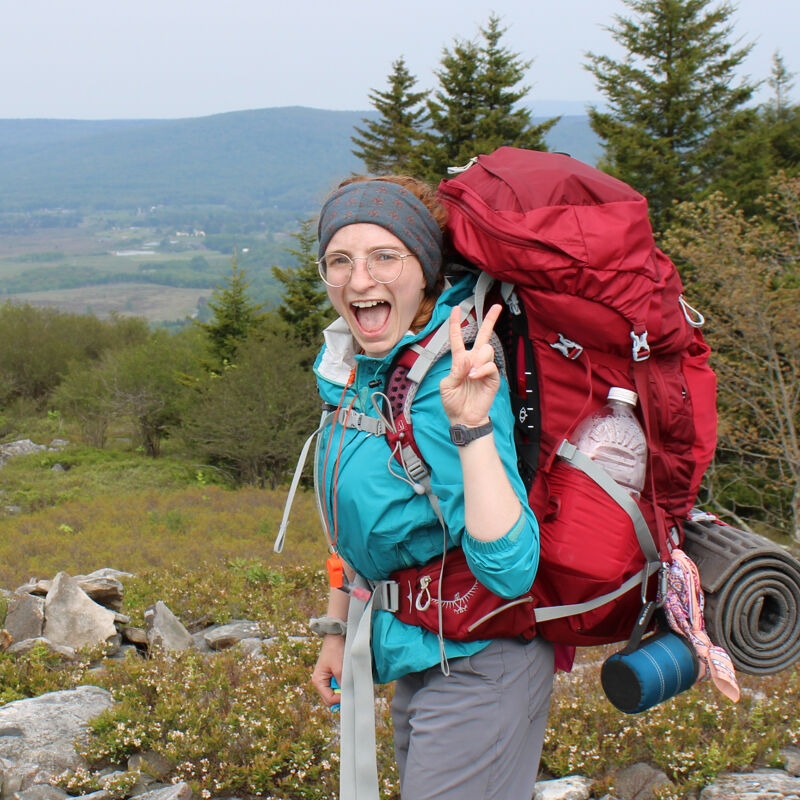 The image shows a woman with a large backpack hiking in a mountainous area. She is wearing a blue jacket, gray pants, and a beanie. She is smiling and holding up a peace sign. The background includes trees, mountains, and a cloudy sky.